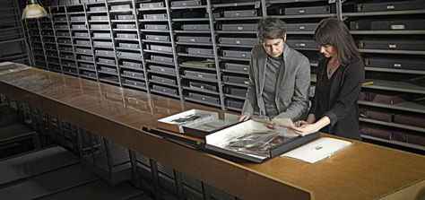AGO Photography curator Sophie Hackett and Vanessa Fleet examine an archival box of negatives in the Art Gallery of Ontario's archives. The archives' shelves are a deep grey, floor to ceiling. The negative box is placed on a long, highly polished wood desk. Hacket matches the archive in a grey cardigan and t-shirt. Fleet wears all black.