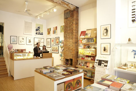 a warm-toned photograph showing Toy Store and gallery Magic Pony's interior. Prints line the walls, and books sit on high display tables. Two people stand behind he cash. The Great Wave off Kanagawa by Hokusai is visible, possibly a modified version where the wave's spray is tiny cartoon rabbits.